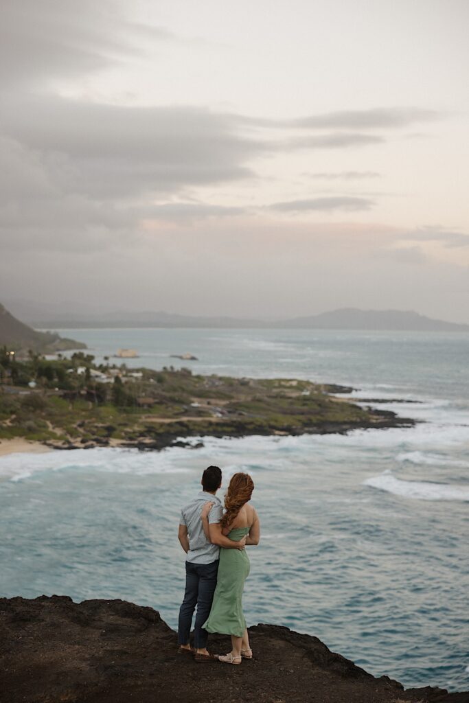 A couple stand atop Makapuu Lookout on Oahu and look out towards the ocean and the island shore