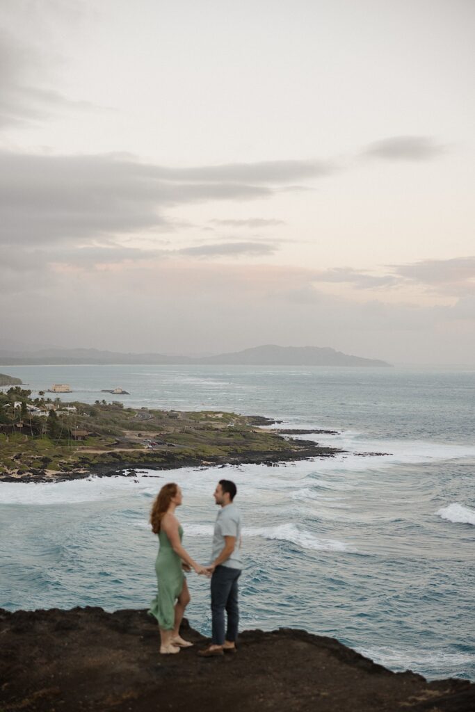 A couple hold hands and face one another atop Makapuu Lookout on Oahu, with the ocean and island's shore in the background
