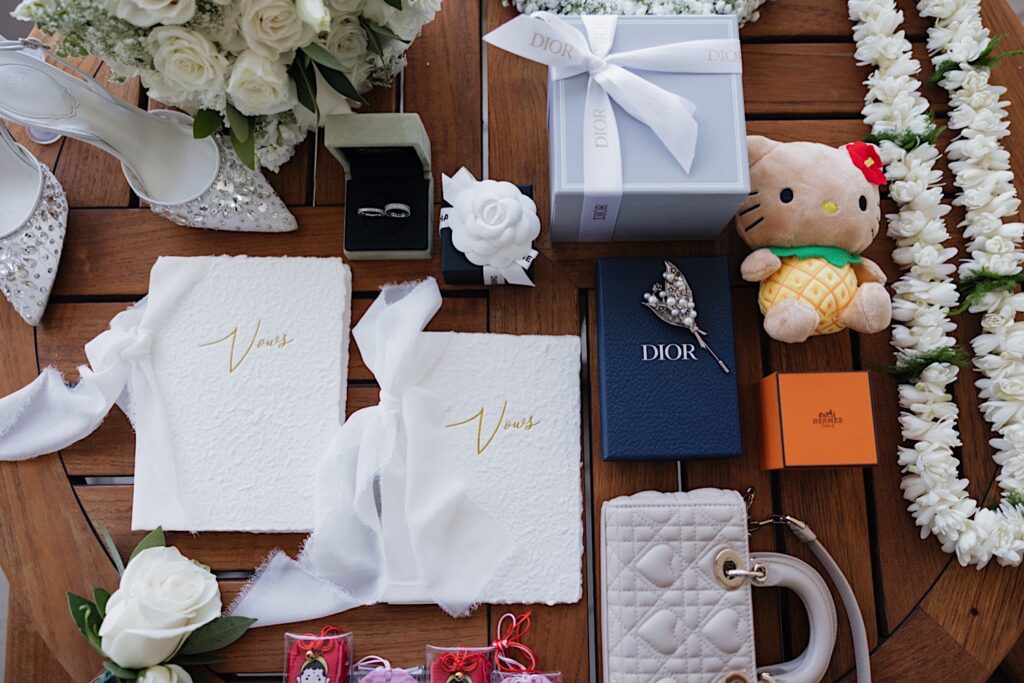 A wedding day flatlay on a wooden table consisting of vow books, rings, shoes, flowers, and more