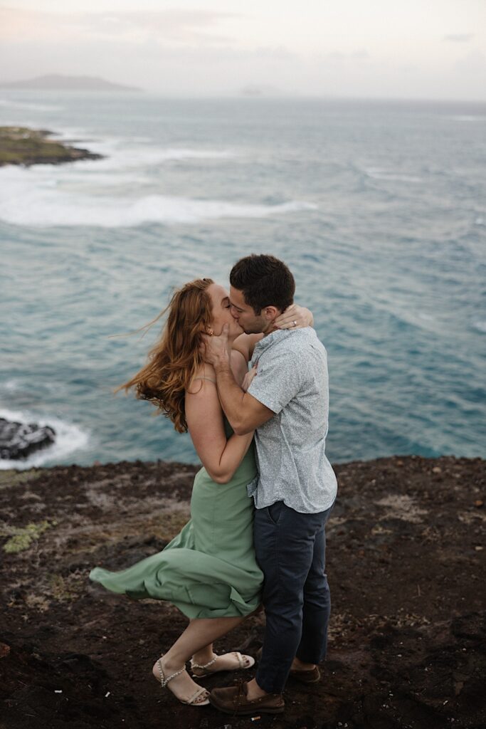 A couple embrace and kiss one another while atop Makapuu Lookout on Oahu with the ocean behind them