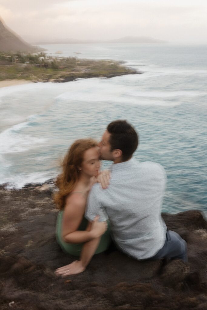 A couple sit together on Makapuu Lookout on Oahu, the man kisses the woman's head as she closes her eyes