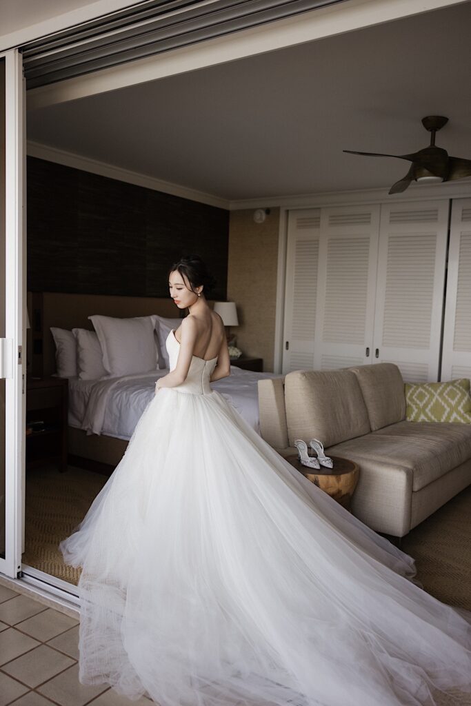 A bride in her wedding dress stands in her hotel room at Four Seasons Oahu in Ko Olina and looks over her shoulder while facing away from the camera