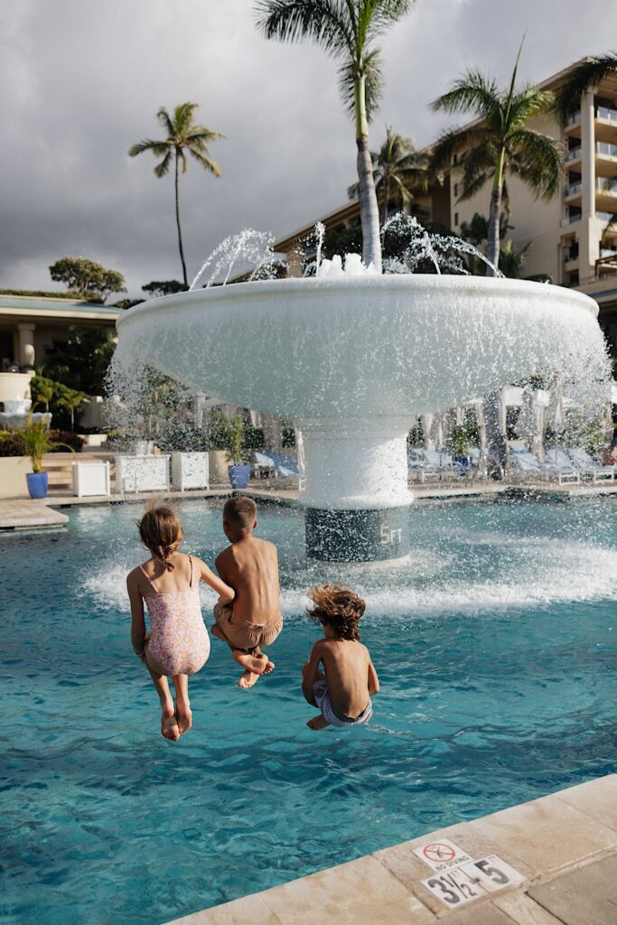 3 children all jump into a pool together at the Four Seasons Resort on Maui, in the middle of the pool is a large fountain