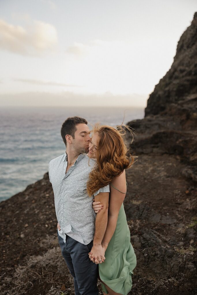 A man kisses a woman's head as she hugs him from behind while the two stand atop Makapuu Lookout on Oahu