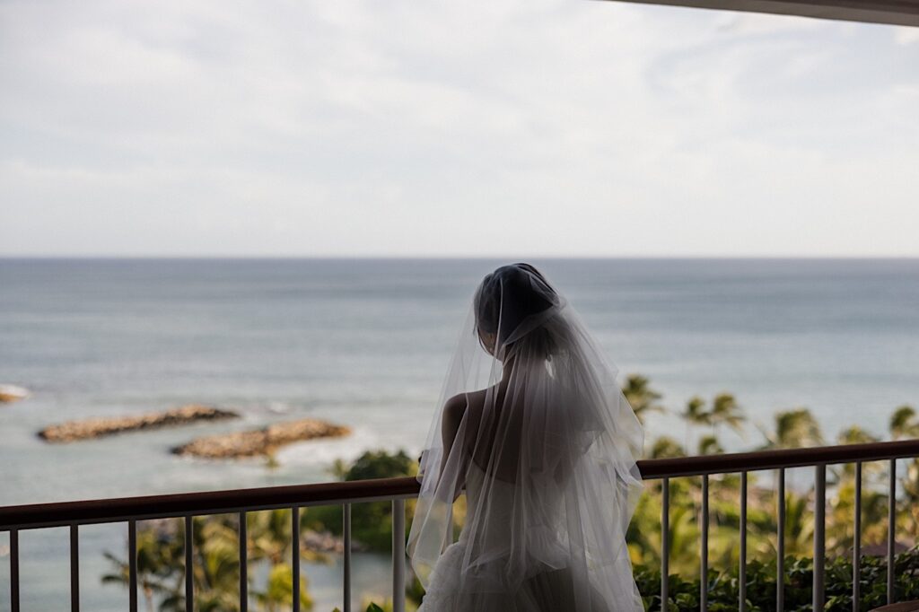 A bride stands and looks out at the ocean from the balcony of her hotel room at the Four Seasons Oahu in Ko Olina before her elopement day