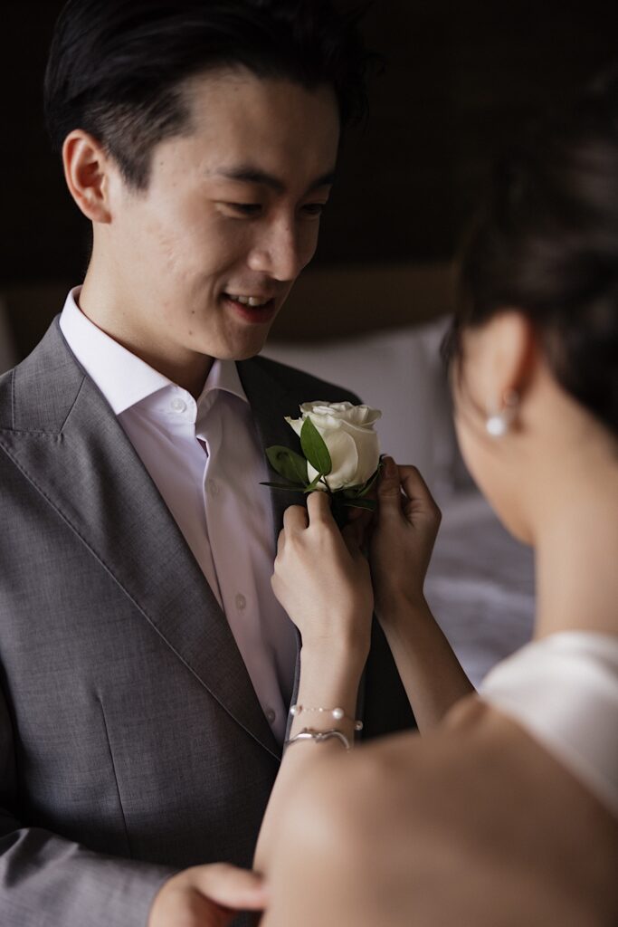 A groom smiles as the bride helps him put on his flower boutonniere while in their hotel room at the Four Seasons Oahu in Ko Olina