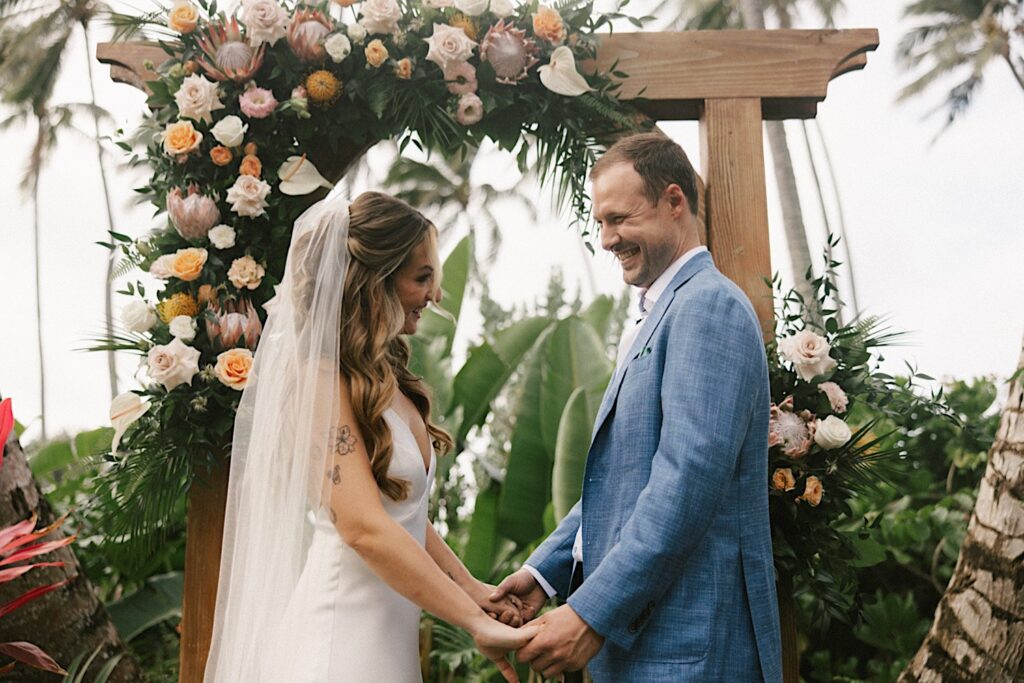 A bride and groom smile while holding hands under a wooden archway decorated with florals during their wedding ceremony at their venue, Male'ana Gardens on Oahu