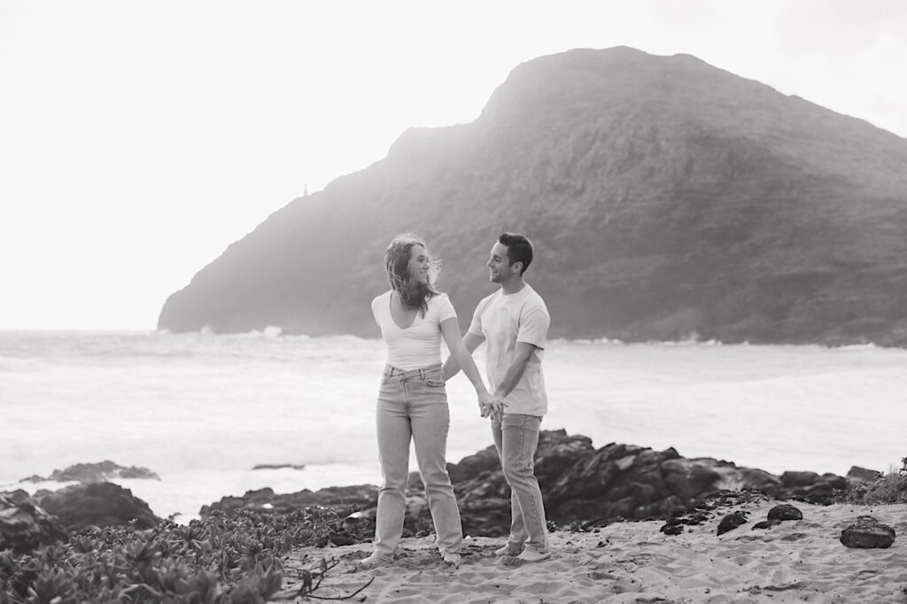 A black and white photo from a romantic honeymoon session where the couple hold hands and look at one another while on the shore of Makapuu Beach of Oahu, Hawaii