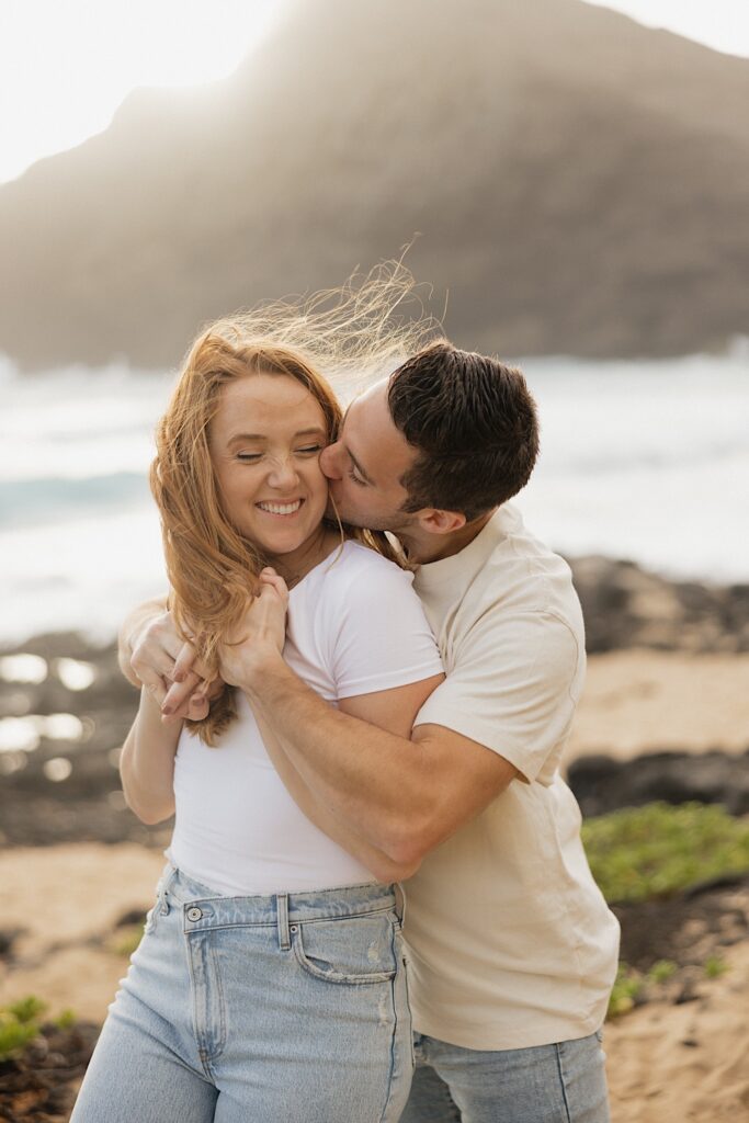 A woman smiles while being kissed on the cheek as a man hugs her from behind during their anniversary session on Makapuu Beach of Oahu