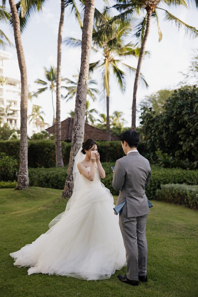 A bride wipes a tear from her eye as the groom reads his private vows to her while they stand near palm trees