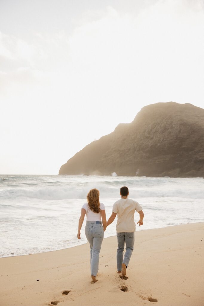 A couple hold hands and run away from the camera towards the waves on Makapuu Beach of Oahu