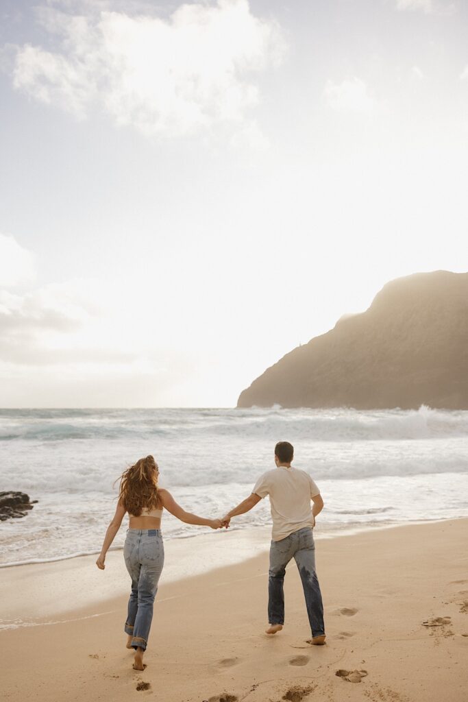 A couple run hand in hand away from the camera towards the ocean waves on Makapuu Beach of Oahu at sunset