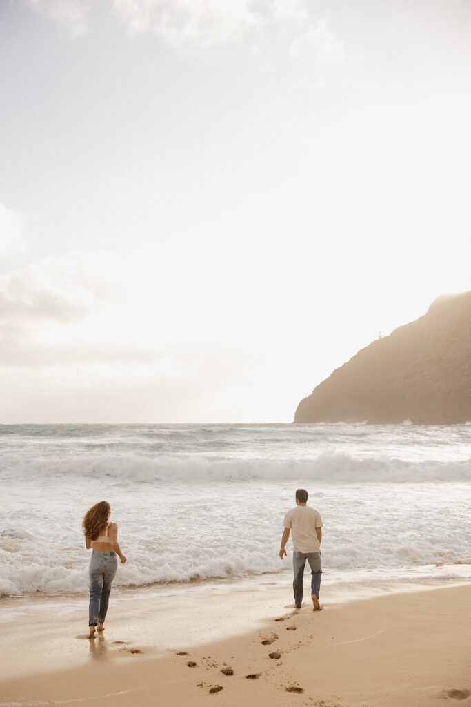 A man and woman both run towards the ocean waves away from the camera while on Makapuu Beach of Oahu