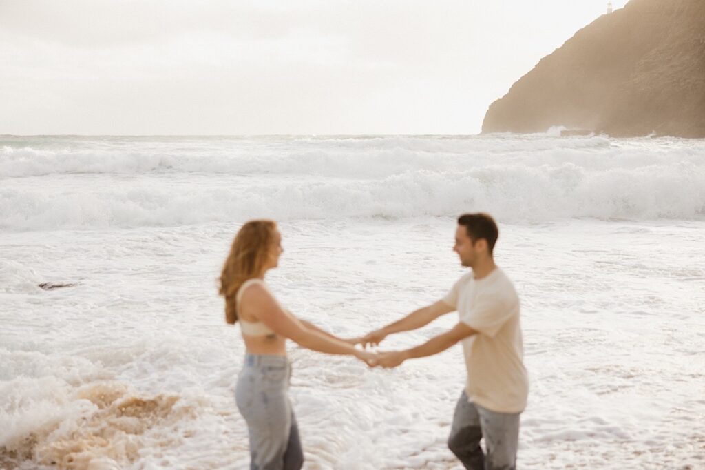 A couple hold hands and face one another while waves crash at their feet during their romantic honeymoon session at Makapuu Beach on Oahu, Hawaii