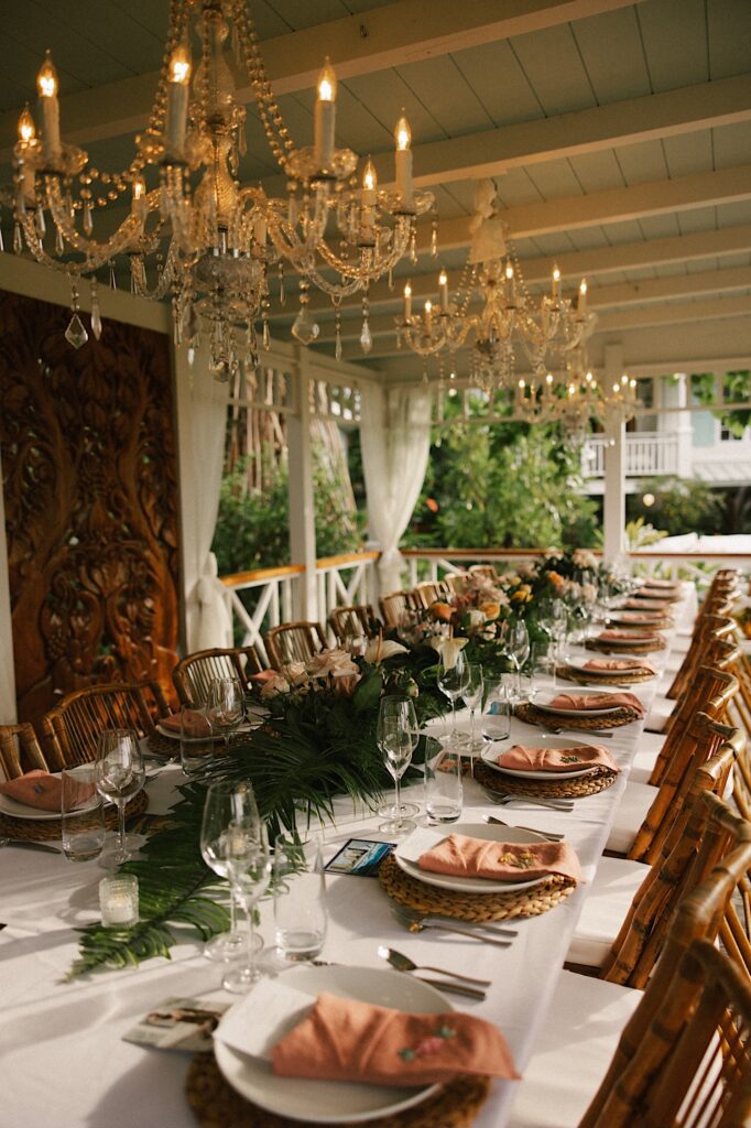 A table is decorated with pastel flowers and sits under a wooden overhang and glass chandeliers, it is the reception space of Male'ana Gardens on Oahu
