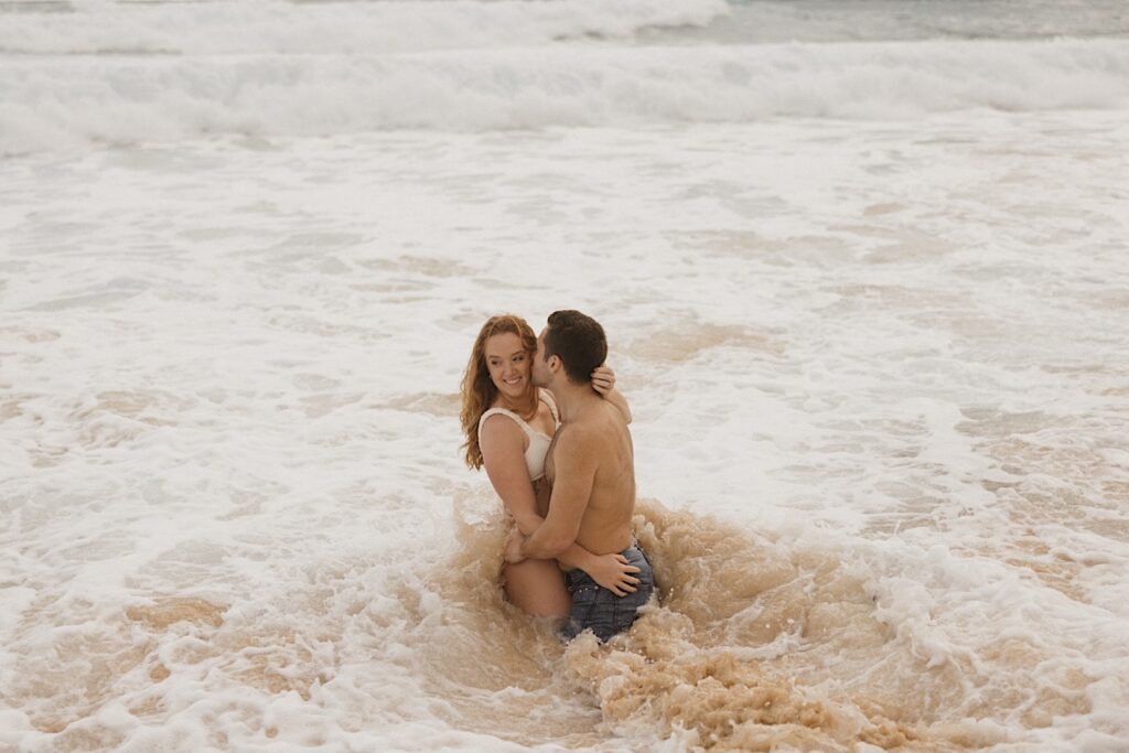 During their romantic honeymoon session, a woman smiles while being hugged and kissed by a man while sitting in the waves crashing onto Makapuu Beach of Oahu, Hawaii