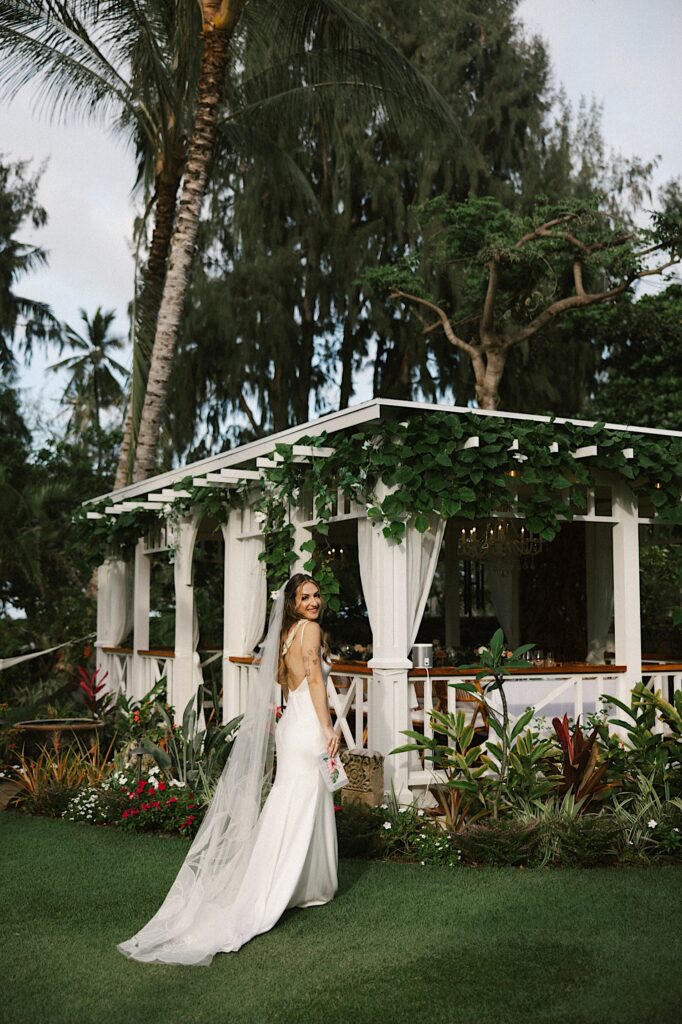 A bride stands next to the reception space at Male'ana Gardens on Oahu and looks over her shoulder to smile at the camera