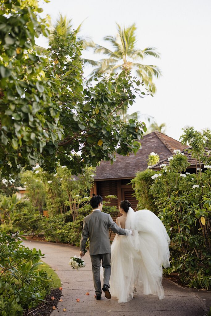 A bride and groom walk along a sidewalk with tropical plants lining it