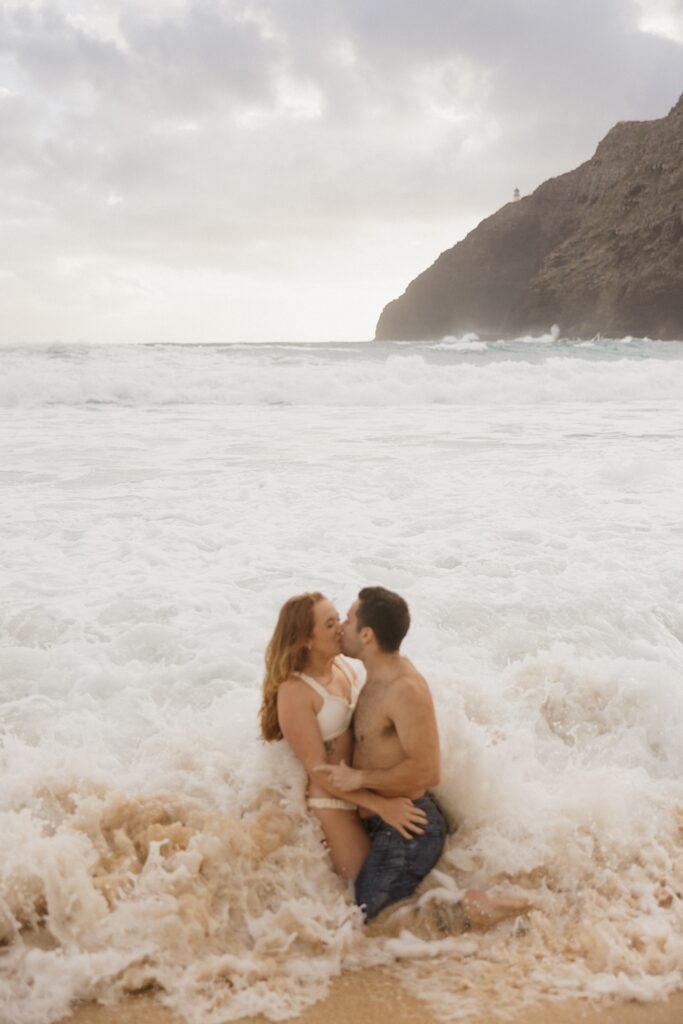 A couple kneel in the sand and kiss as waves crash around them while on Makapuu Beach of Oahu
