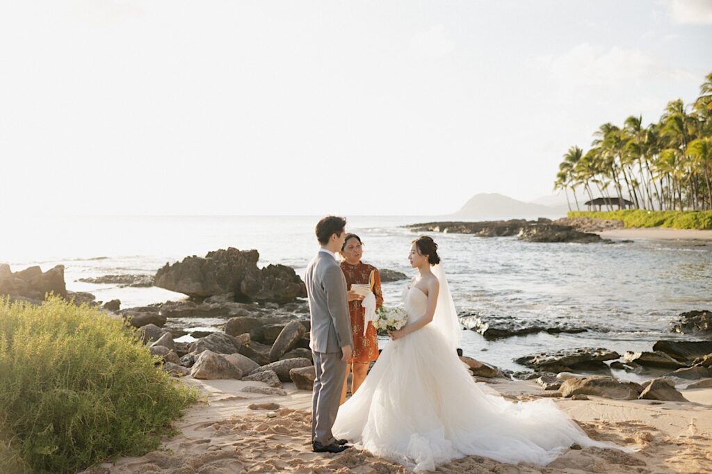 A bride and groom stand facing one another on a beach near the Four Seasons Oahu in Ko Olina as the officiant of their elopement ceremony speaks to them