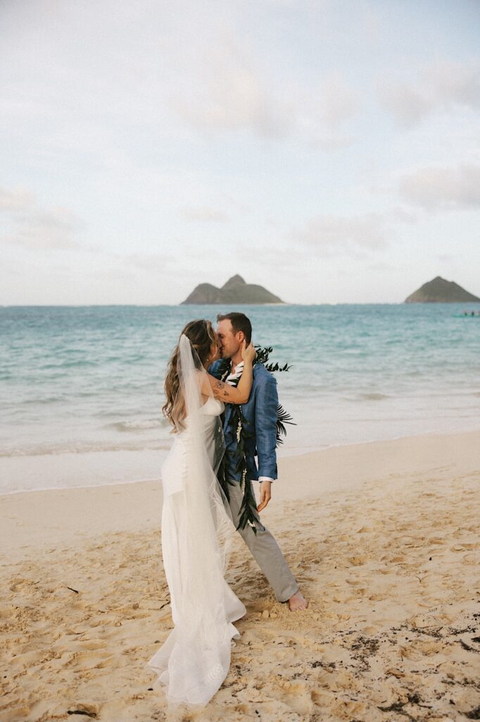 A bride and groom kiss one another while on a beach of Oahu near Male'ana Gardens with the ocean and some smaller islands in the background