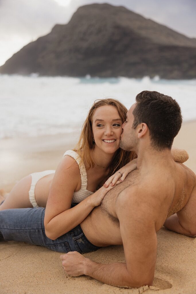 A woman smiles while being kissed by a man as she lays on top of him while on Makapuu Beach of Oahu
