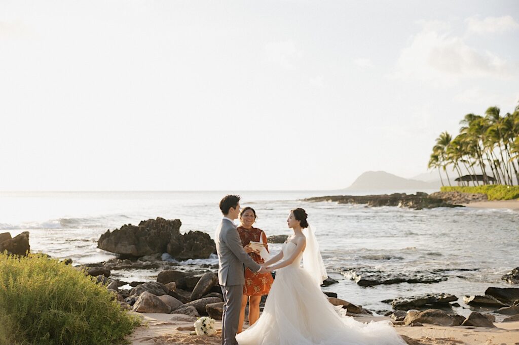 A bride and groom hold hands while facing each other on a beach near the Four Seasons Oahu in Ko Olina during their elopement ceremony