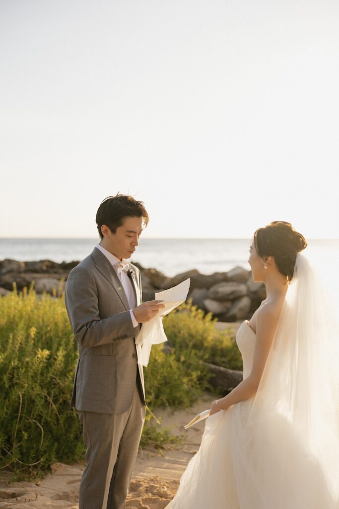 A groom reads his vows to the bride while the two stand on a beach in Oahu as the sun sets behind them
