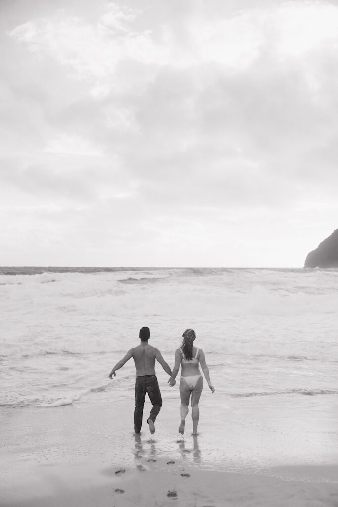 Black and white photo of a couple running into the ocean together away from the camera while on Makapuu Beach of Oahu