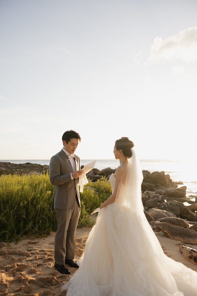A groom reads his vows to the bride as she listens while the two stand on a beach in Oahu as the sun sets