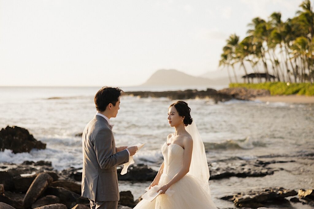 A bride listens as the groom reads his vows to her while they stand on a beach near the Four Seasons Oahu in Ko Olina for their elopement ceremony