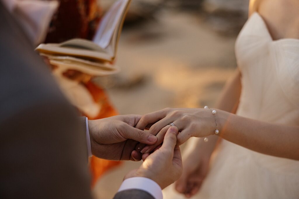 Close up photo of a groom holding the bride's hand as he puts her wedding ring on during their elopement on a beach near the Four Seasons Oahu in Ko Olina