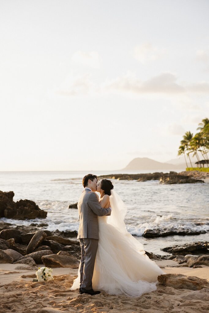 A bride and groom kiss one another while standing on a beach of Oahu while the sun sets behind them after their elopement
