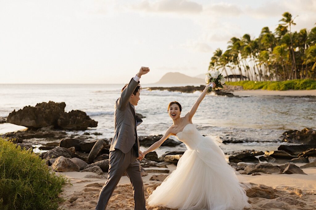 A bride and groom celebrate and raise their arms in the air while on a beach near the Four Seasons Oahu at Ko Olina after finishing their elopement ceremony