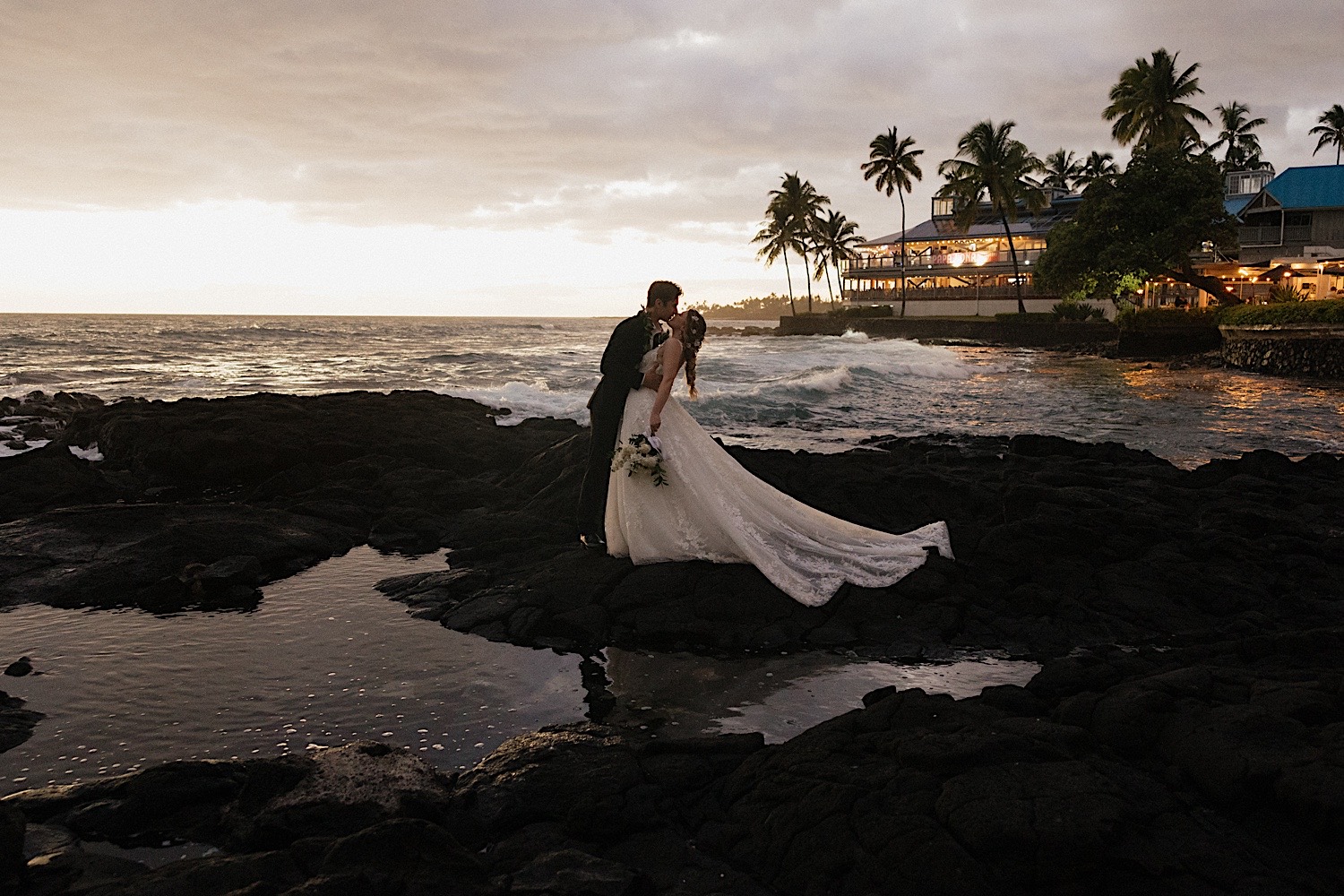 A bride and groom kiss one another while on a rocky shore in Hawaii with their wedding venue in the background behind them