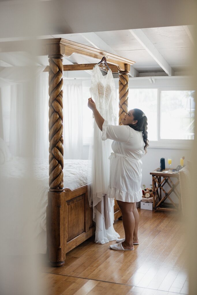 A bride smiles while looking at her wedding dress that's hanging from a wooden bed frame