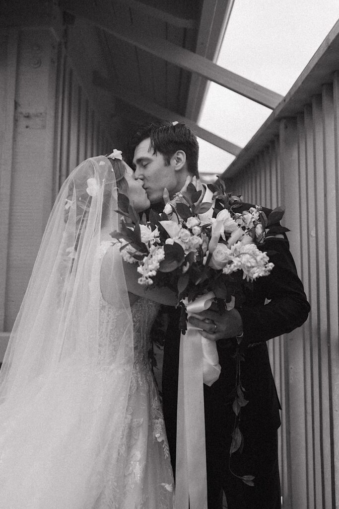 Black and white photo of a bride and groom kissing one another while under an overhang