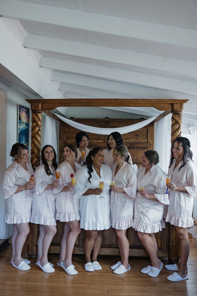 A bride and her 8 bridesmaids all smile while holding mimosas before getting ready for the wedding day