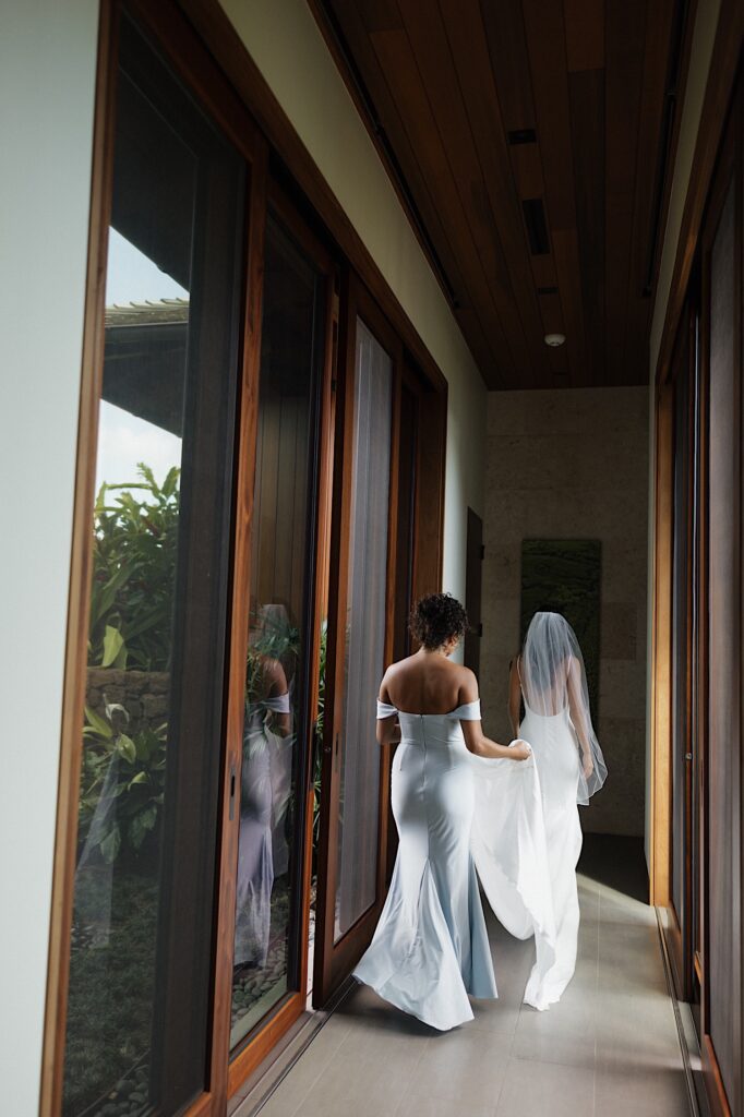 A bride walks out of her getting ready space as a bridesmaid helps with the tail of her dress