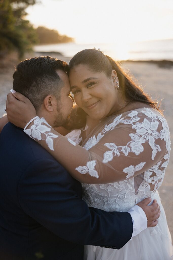 A bride smiles at the camera while hugging the groom as they take portrait photos on the beach at sunset