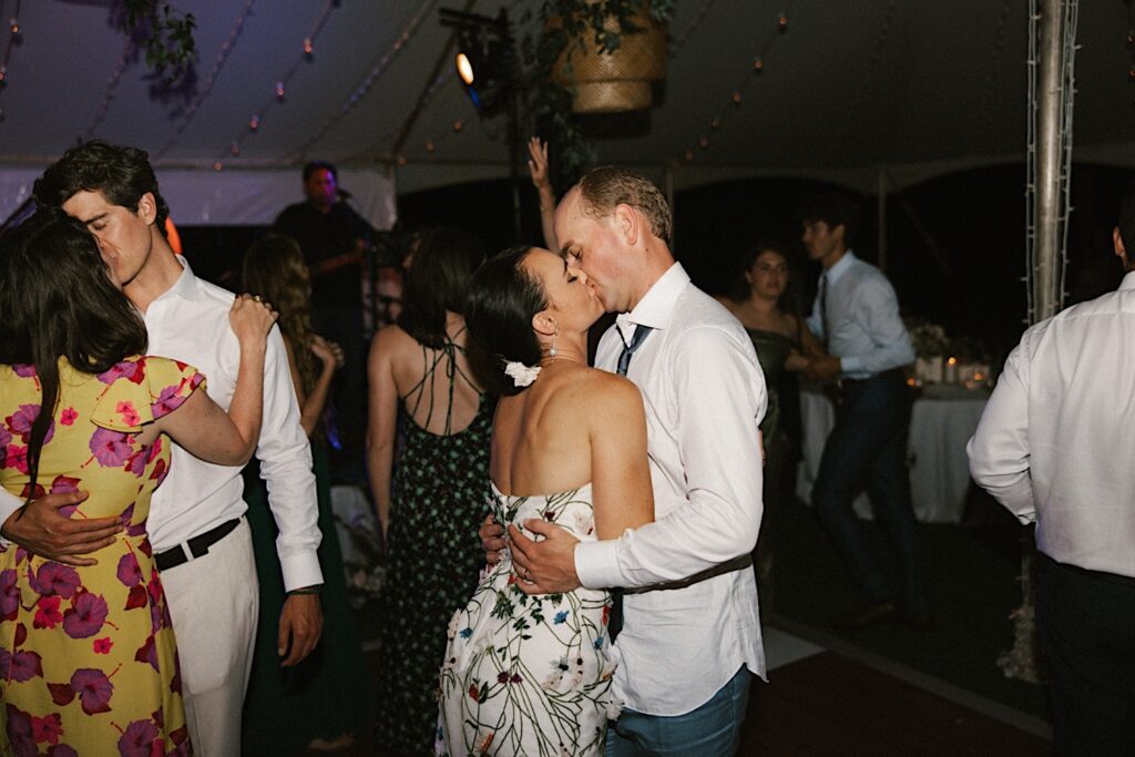 A bride and groom kiss one another during their wedding reception under a tent in Hawaii as guests dance around them