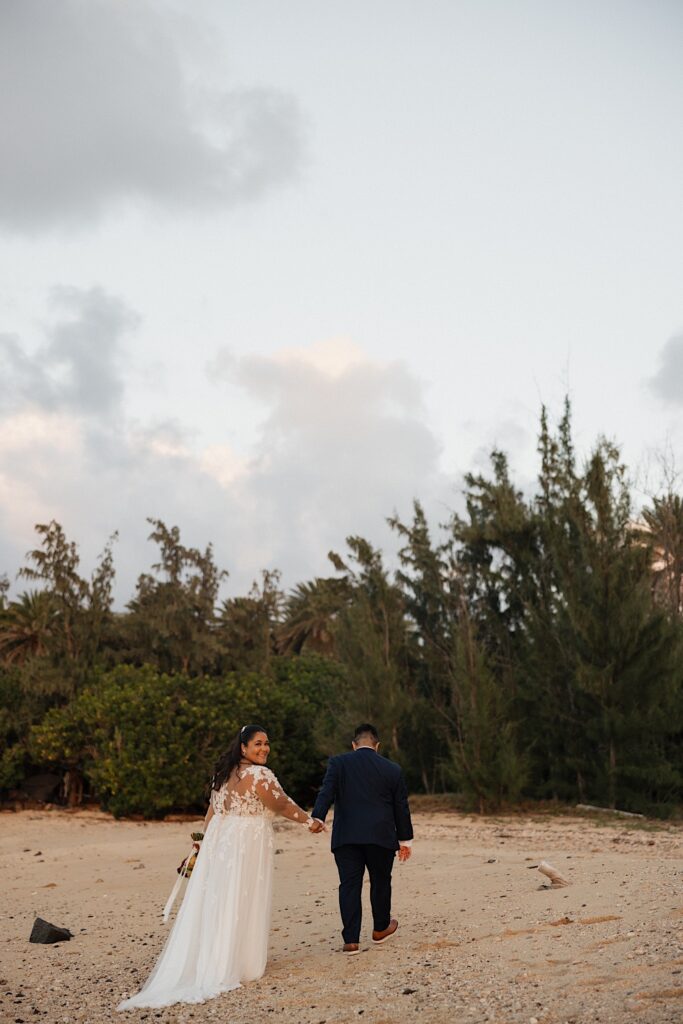 A bride smiles over her shoulder towards the camera while walking away with the groom on a beach of Oahu