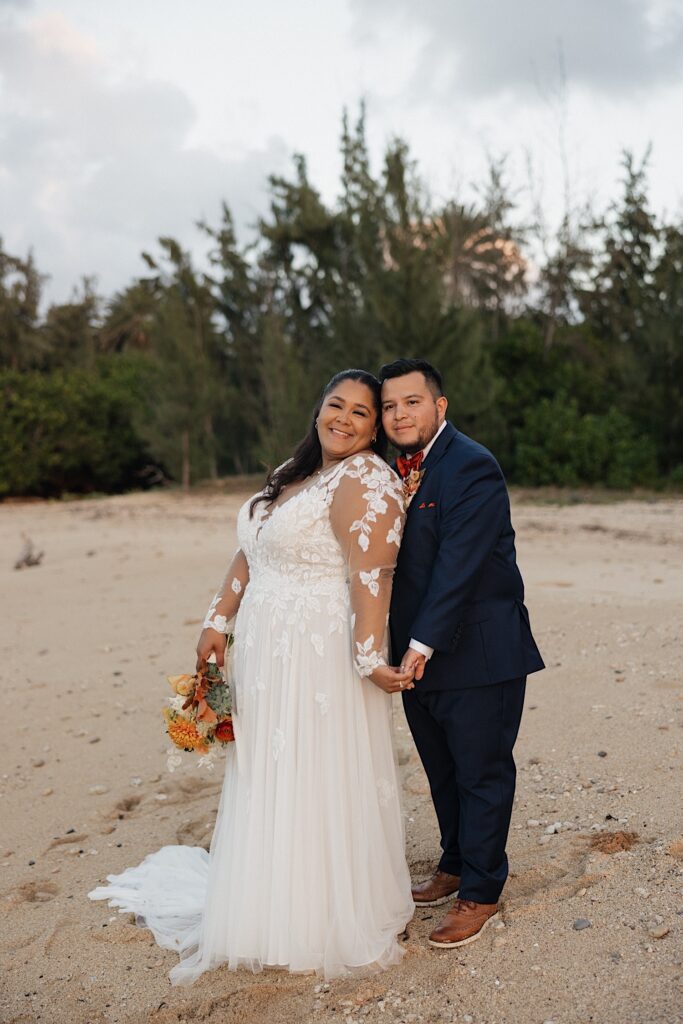 A bride and groom smile at the camera while holding hands and standing on a beach of Oahu