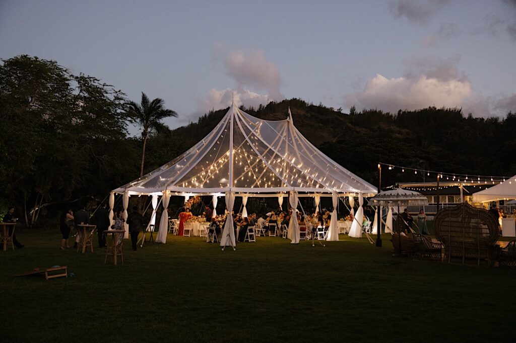 A clear tent is lit up with string lights during an outdoor wedding reception at a villa on Oahu