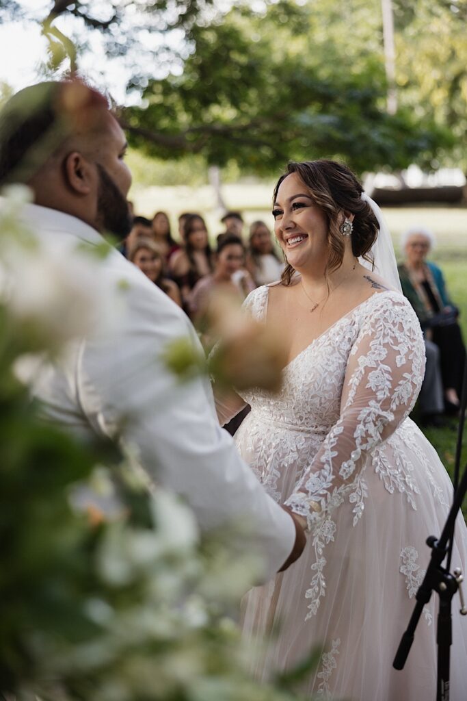 A bride smiles while holding the hands of her groom during their outdoor wedding ceremony