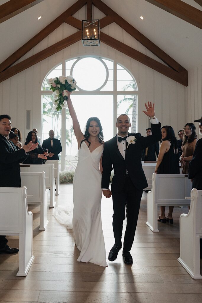 A bride and groom hold hands and raise their arms in the air as they exit their wedding ceremony together