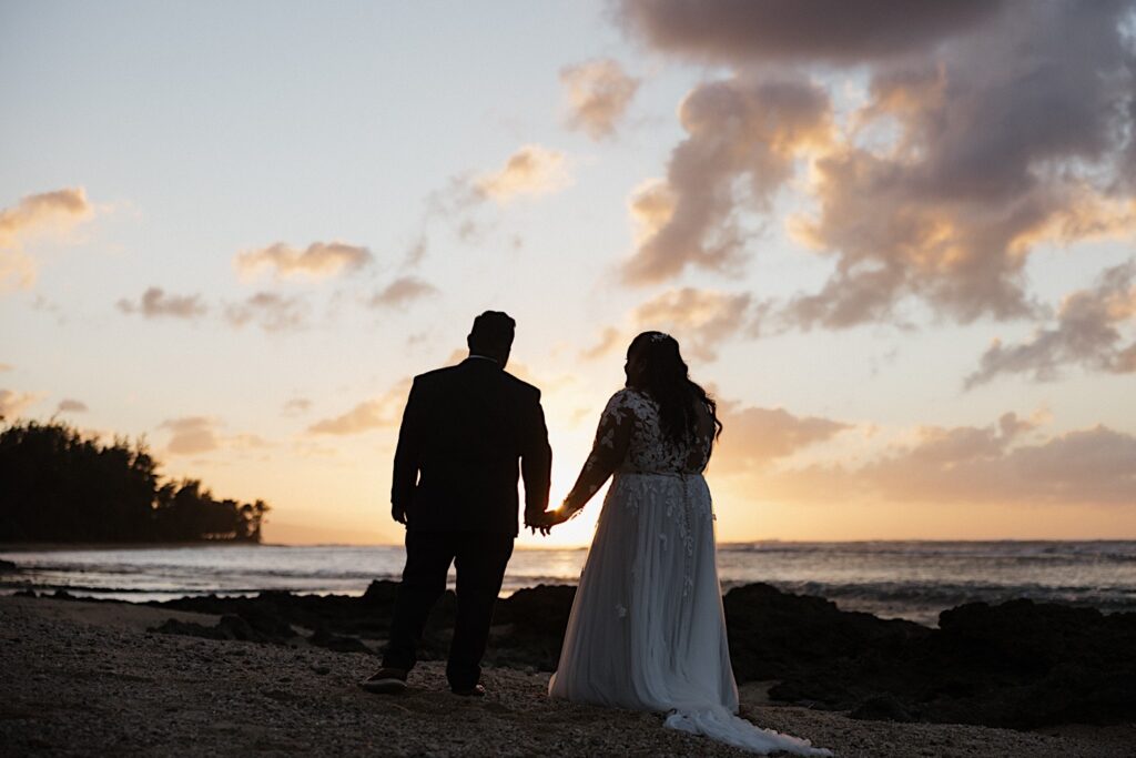 A bride and groom stand together on a beach in Hawaii and hold hands as they look out towards the sun setting on the ocean