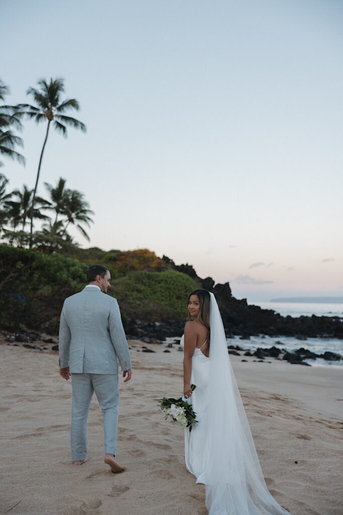 Bride in white dress looks back at camera while walking with Groom in a grey suit while walking along a beach during their Maui Elopement. 