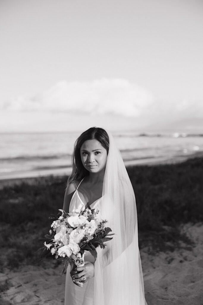 Black and White photo of bride wearing silky white dress holding floral arrangement on beautiful Maui beach in Hawaii after elopement ceremony. 