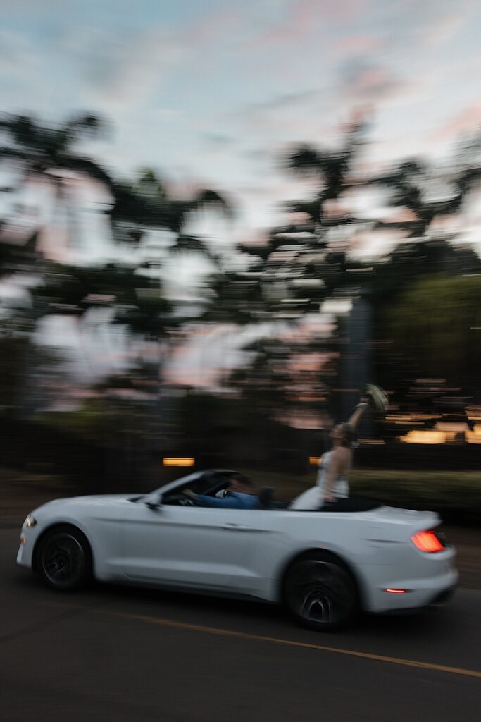 Bride sits on the back of convertible holding her bouquet in the air as Groom drives the car into the sunset after wedding ceremony in Maui. 